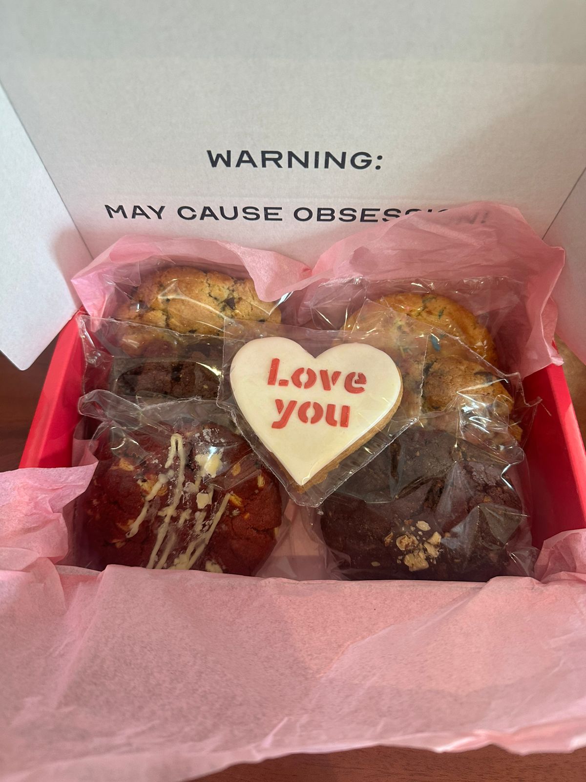 Box of assorted cookies with a heart-shaped 'Love you' cookie and warning label.